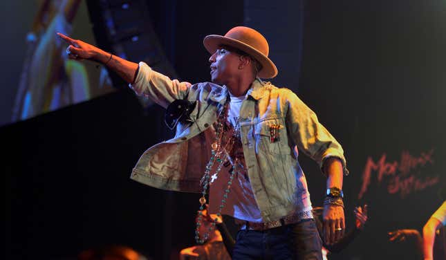 Pharrell Williams on stage in a rimmed hat, a denim jacker and many necklaces. 