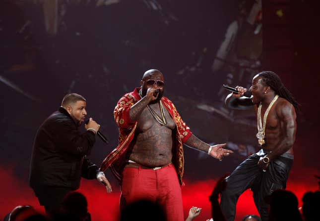 Rick Ross stands in between DJ Khaled and Ace Hood, wearing red pants, and an open shirt. 